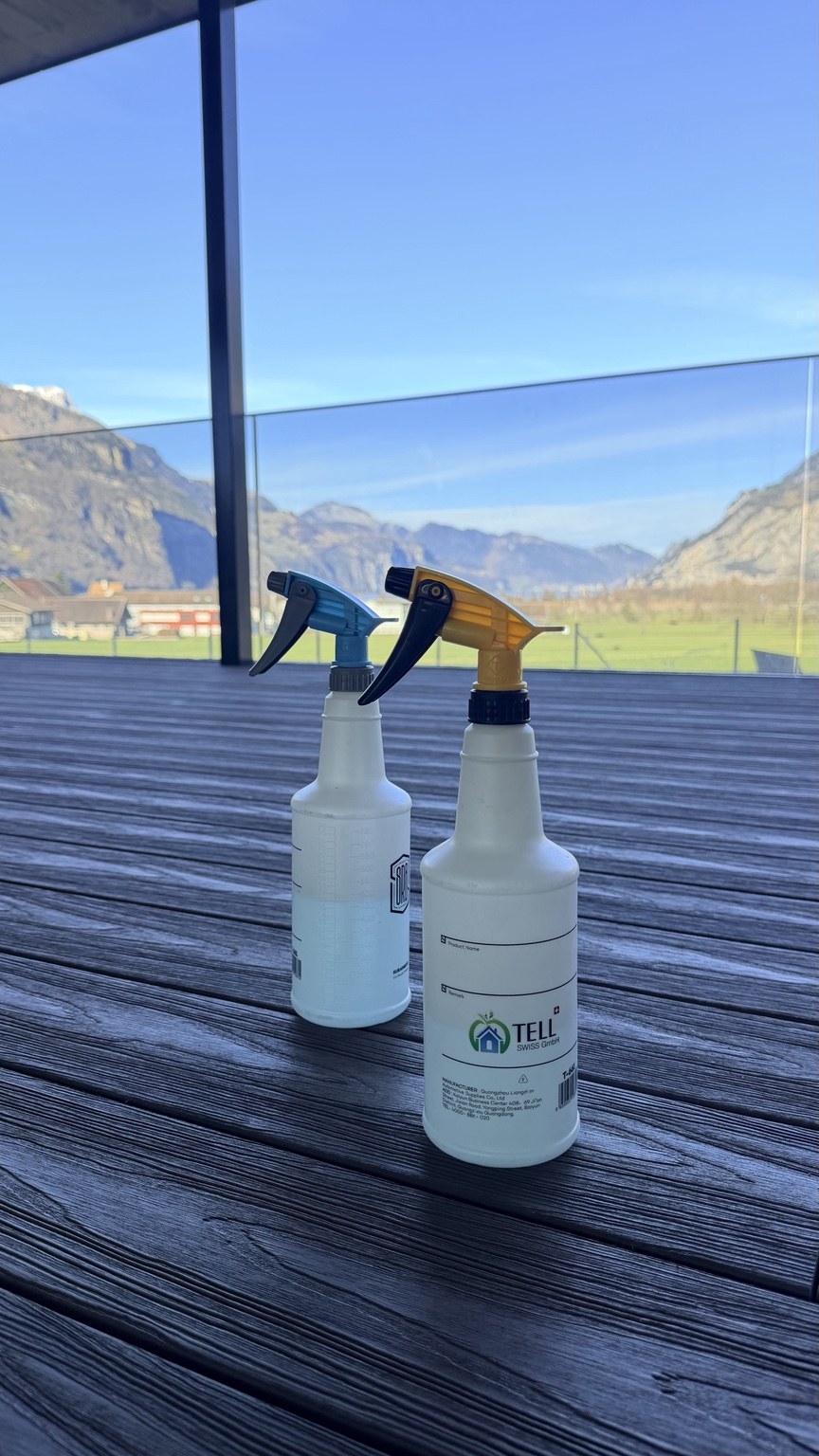 Zwei Reinigungssprühflaschen, eine mit Tell Swiss Logo, auf einer Holzterrasse vor einem Glasgeländer und Bergpanorama